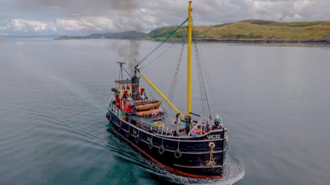 The Clyde Puffer SL VIC 32 - a small steamboat in dark blue/black, with a yellow mast, sails through water.