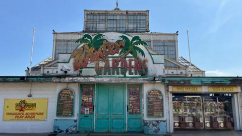 Great Yarmouth's winter gardens, with an illuminated sign depicting palm trees, on the façade of the Victorian structure which is white, and made from cast iron and glass, much of which is boarded up on the inside. On the façade are other signs and some arcade machines on the right of the image.