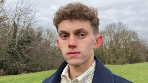 Man stares at the camera with a blank expression. He is stood in a field with bare branches lining a hedgerow. He wears a checked shirt, dark sweatshirt and thick navy blue coat.