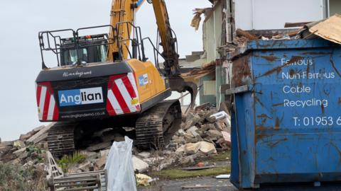 A close-up of a digger demolishing a home. A large industrial blue skip is on the right. Rubble and debris from the home is scattered along the ground and underneath the digger.