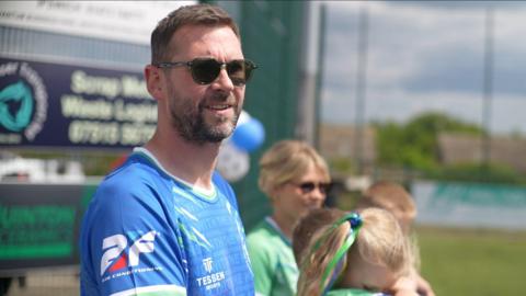 A side-on view of a man wearing a blue football top and sunglasses, standing on the sidelines of a football pitch. He has brown hair and a beard. To his left is a woman with blonde hair, sunglasses and a green football top, a girl with blonde hair and a green football top, and the top of the heads of two other children.