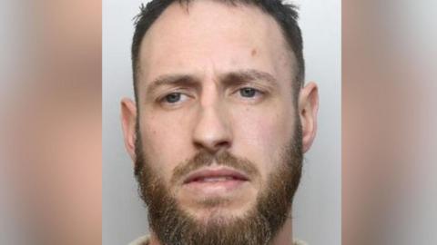 A mugshot of a man with blue eyes, a moustache and beard
