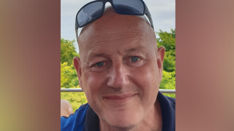 Jeff Blair with sunglasses on his head and wearing a blue top. He is bald and is smiling in the close-up shot. There are trees behind him.