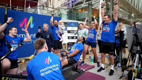 A group of people wearing blue shirts reading "Thanks a Million" on the front and "momentum fitness charity" on the back. A man is in the middle of the group sat on a static rowing machine.
