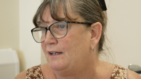 A head and shoulders view of Angela, wearing glasses with tied-back greying hair and hoop earrings