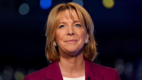 Hazel Irvine is pictured in a burgundy suit jacket with a white top underneath. She is smiling and the background is dark and blurred. 