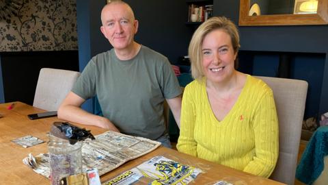 Phil Betts, in a green t-shirt, and Charlotte Betts, wearing a yellow jumper,  sit together at a table with contents of the time capsule on top, including a newspaper and a glass jar  