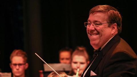 Andrew White with brown hair and glasses wearing a black suit smiling as he conducts the Youth Concert Band with a baton and some musicians playing instruments in the background