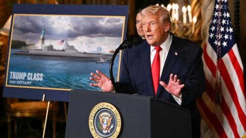 US President Donald Trump stood speaking at a lectern in a navy suit with red tie next to an image of a tanker