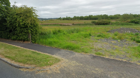 A view into a field on Ridge Avenue, Burnley. The field has no gate and leads straight from the road. The grass is short in places, and there are shrubs and trees in the field.