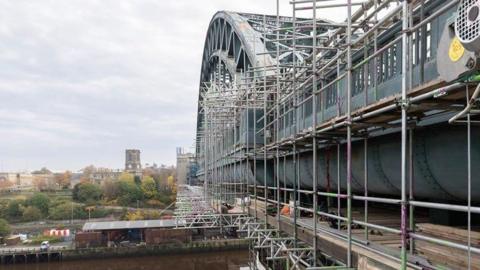 To the right, there is the large green metal arched Tyne Bridge stretching over the river, with scaffolding covering it. In the distance there are trees and buildings and a car park. It is a cloudy day.