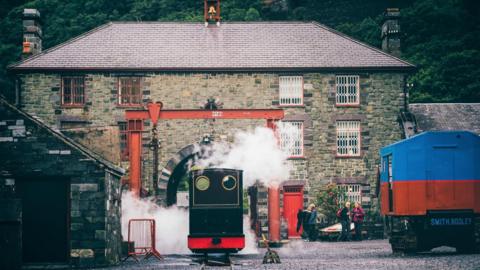 Old buildings and machinery with the back of a red steam locomotive visible in the centre of the shot as visitors look on