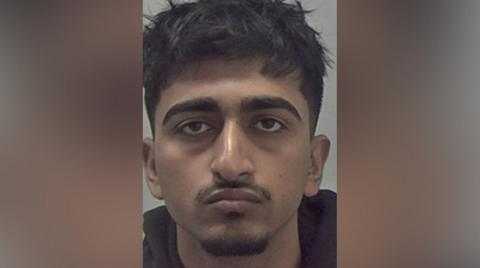 A mugshot of a man with messy black hair and a moustache and goatee beard. He is wearing a black top.