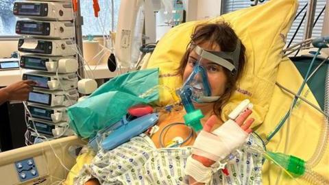 A woman with brown hair in a hospital bed with multiple tubes attached to her body.