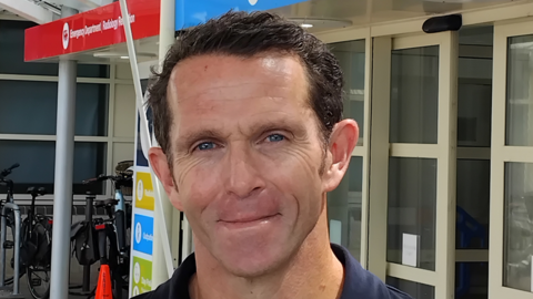 A man looking into the camera and smiling. He has brown hair and is wearing a navy shirt. He's standing in front of the entrance of a hospital.