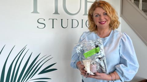 Kerry smiles at the camera while holding the plastic bag containing a brush and two smaller bottles. She stands in front of a white wall with dark green image of a leaf and the word STUDIO behind her. She has chin length wavy blonde hair and wears a blue blouse.