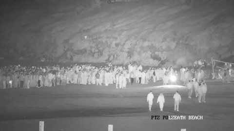 A black and white image shows hundreds of young people gathered together on a beach. 