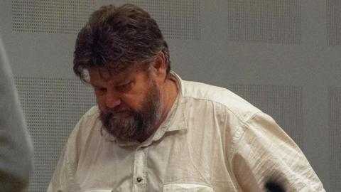 Carl Beech, a man in his 50s with dark grey hair and a mid-length beard, walking along with his head down, wearing a cream-coloured shirt.