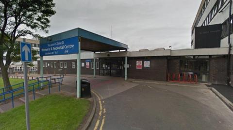 Exterior of the neonatal department at New Cross Hospital. A single-storey, flat-roofed building is in the foreground with an NHS sign at the front