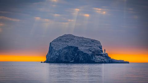 The Bass Rock in the sea. The sky is dark with yellow light poking through the clouds. The horizon is a bright orange colour.