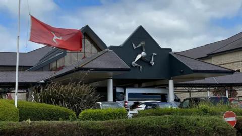 The entrance to Noble's Hospital, which has an angled roof over pillars with a metal three legs of Mann sculpture on the front. A row of cars is parked behind a row of hedges. A red Manx flag is flying on a flagpole on the left.