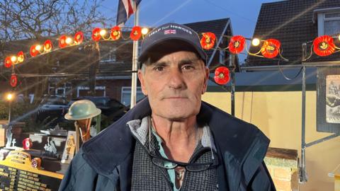 A head and shoulder photo of a man looking directly into the camera. He is wearing a navy Help for Heroes cap and has glasses dangling from his neck. He is wearing a grey quarter zip and a navy coat. He is stood in front of his display. Poppies are on the archway behind him and there are lights. 