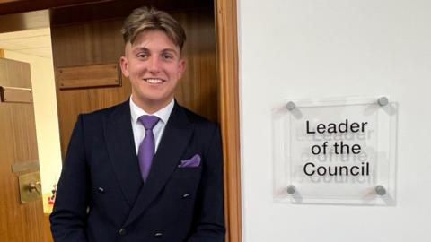 George Finch leans against a door frame. A sign to his left reads 'leader of the council'. He wears a dark suit, white shirt and purple tie.