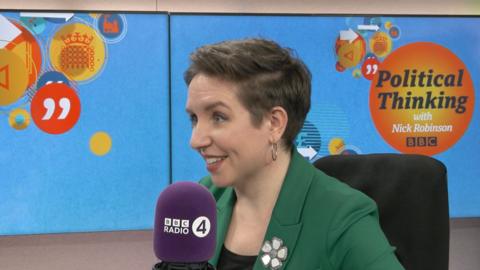 Carla Denyer, wearing a green suit, sitting and smiling in a radio studio in front of a BBC Radio 4 microphone