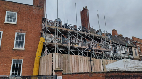 A black and white timbered building covered in scaffolding and surrounded by wooden fencing with a red brick building alongside