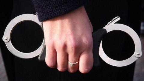 A female police officer's hand, with a silver ring on the middle finger, holds a pair of metal handcuffs