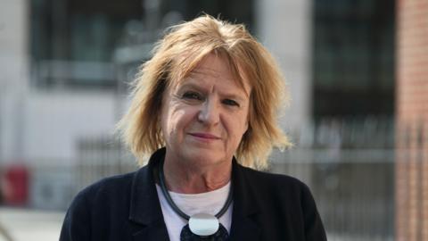 Head and shoulders photo of Deborah Coles. She has straight blonde shoulder-length hair with a centre parting. She is wearing a dark jacket with a white top underneath, and a large black and silver necklace. She is standing in front of a building with railings outside, and is looking directly into the camera