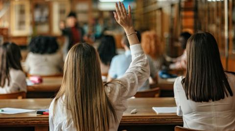 Students sitting in a classroom. The photo is taken from behind them. One of the pupils has their hand raised.