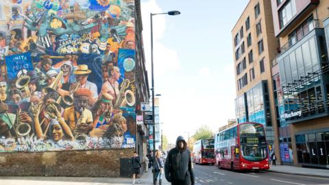 A mural on a wall with colourful drawings of musicians and local people. It stands opposite a modern Hackney council building. 