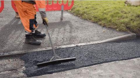 A person is working on repairing a road. They are visible from the hips down, wearing high visibility orange clothing, yellow gloves and dark work boots. There is a bright orange temporary barrier behind them, and they're using a long handled tool to level out the new tarmac that has been laid. There is a grassy area to the right of the works.