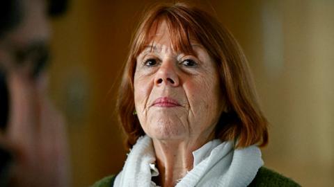 A woman wearing a grey jacket and scarf looks directly at the camera expressionless.