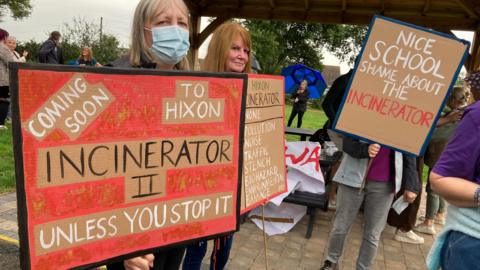Residents gather at a protest. Some are holding placards and signs opposing the plans for the clinical waste incinerator. 