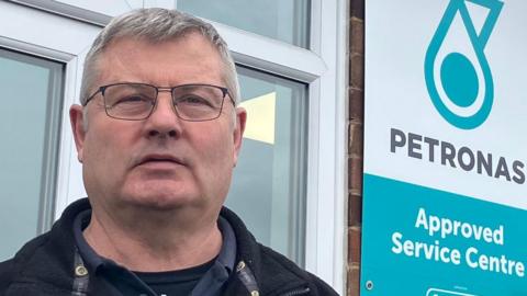 Dave is standing outside his business premises, which has windows and a brick wall. He is wearing a dark jacket and dark clothing underneath. He is also wearing glasses. He has short, grey hair.
