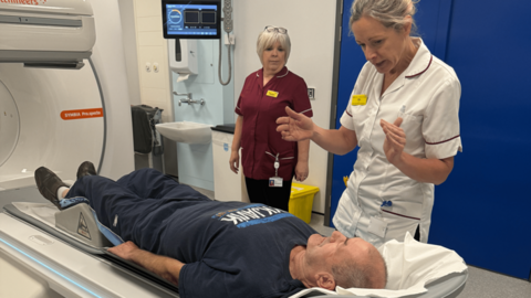 Two women in medical uniform stand by a man lying on a scanner unit.