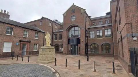 A general street view of the front of the Carlisle Crown Court red brick building. It has the royal coat of arms above the front door, there is a statue outside and the street is a mix of cobbled and paved.