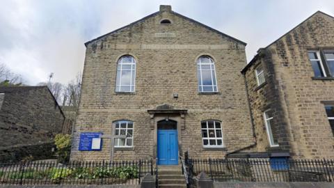 A square stone building with a blue door, four windows and Methodist Church inscribed on the front in stone. A blue sign on the left hand side, a cross standing in the flowerbed in front of the building. 