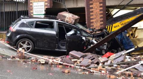 A crashed black Seat Leon car embedded in the side of the St John's car park. The vehicle is surrounded by rubble and brickwork following the collision.