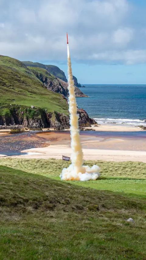 Rocket launches from grass-covered land near a beach by the sea