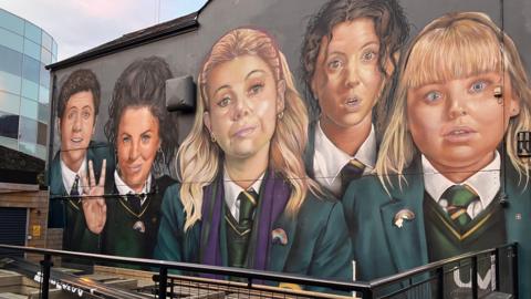 The Derry Girls mural in Derry, featuring four of the characters from the TV show. They are dressed in school uniforms. In the foreground is a seating area while the awning that partially covered the mural has now been removed