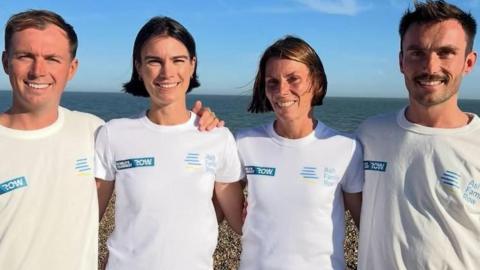 Two men and two women stand on a pebbled beach wearing matching white “World’s Toughest Row – Ash Family Row” T‑shirts, with the sea and blue sky behind them.