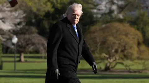 Donald Trump, wearing a dark jacket, looks at the camera while walking on a lawn.