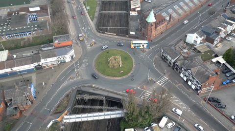 Overhead drone view of the green centre of the roundabout above the rail line. It's surrounded by vehicles driven on the roads and buildings.