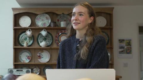 Lucy is stood at a dining table, her laptop in front of her. She wears a dark blue cardigan. Shelves of crockery are behind her.