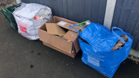 A green food caddy next to recycling, placed on the pavement in white and blue bags and a cardboard box.