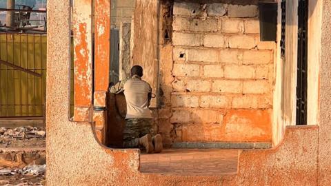 A man sits on his knees in a brick building looking through a window