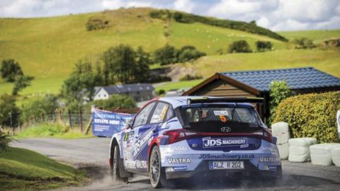 Rally car driving on a road past fields of grass